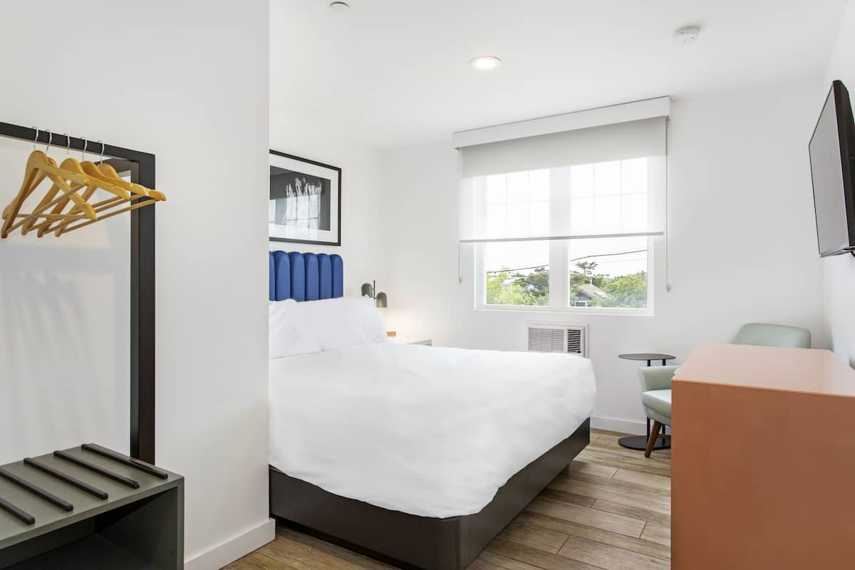 A modern hotel room with a bed, a desk, a chair, a window with blinds, a radiator, and a wall-mounted TV.
