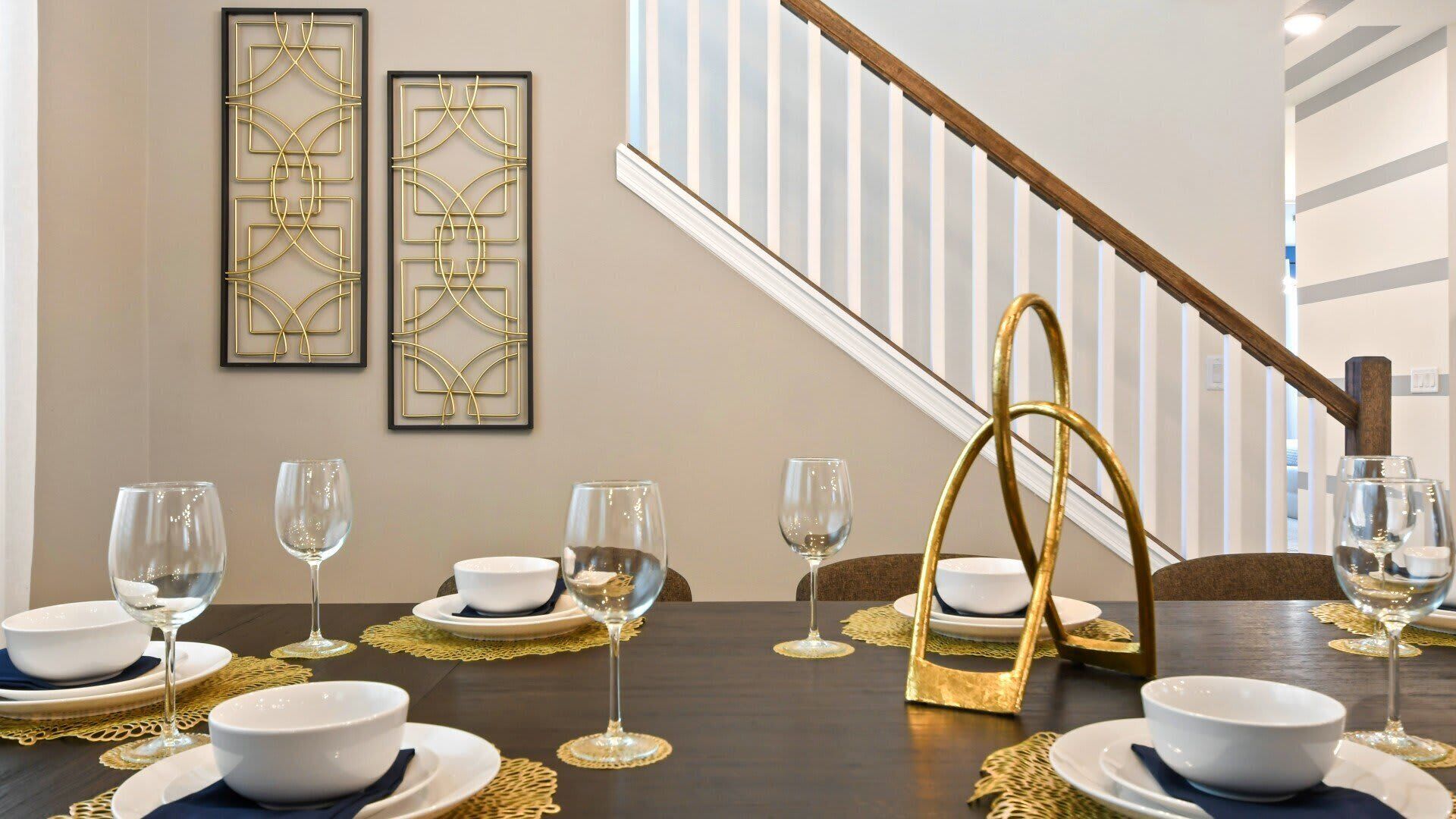 A dining area with a dark wooden table set for a meal, gold placemats, and white dinnerware. Two framed abstract gold wall art pieces are visible above the table.
