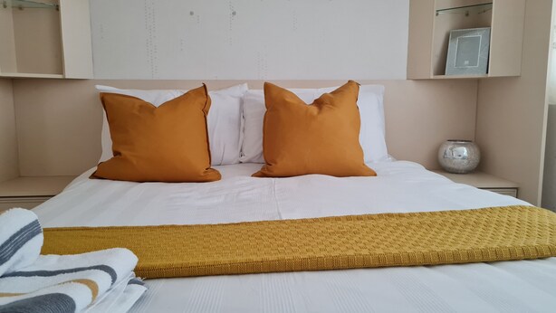 Una cama bien hecha con ropa de cama blanca y color mostaza, dos almohadas color mostaza y un edredón color mostaza.
