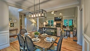 A spacious dining area with a wooden table set for six, surrounded by chairs. A modern chandelier hangs above, and a staircase leads to an upper level. - Lakefront family home w/ private 2-story dock & NEW outdoor deck for dining! (Dawsonville)