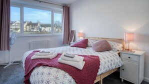 Ein ordentlich bezogenes Bett mit einem blumigen Überwurf, einer rosa Strickdecke und einem Holzkopfstück. Zwei Nachttischlampen, ein weißer Nachttisch und ein Fenster mit Vorhängen sind ebenfalls vorhanden.