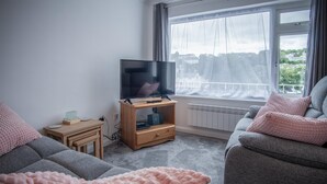 Ein Wohnzimmer mit Fernseher auf Holzfuß, einer Couch und einem Fenster mit Blick auf Gebäude.