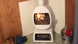 A wood-burning stove with a white chimney, a shelf with plants, and a pair of boots on the floor.