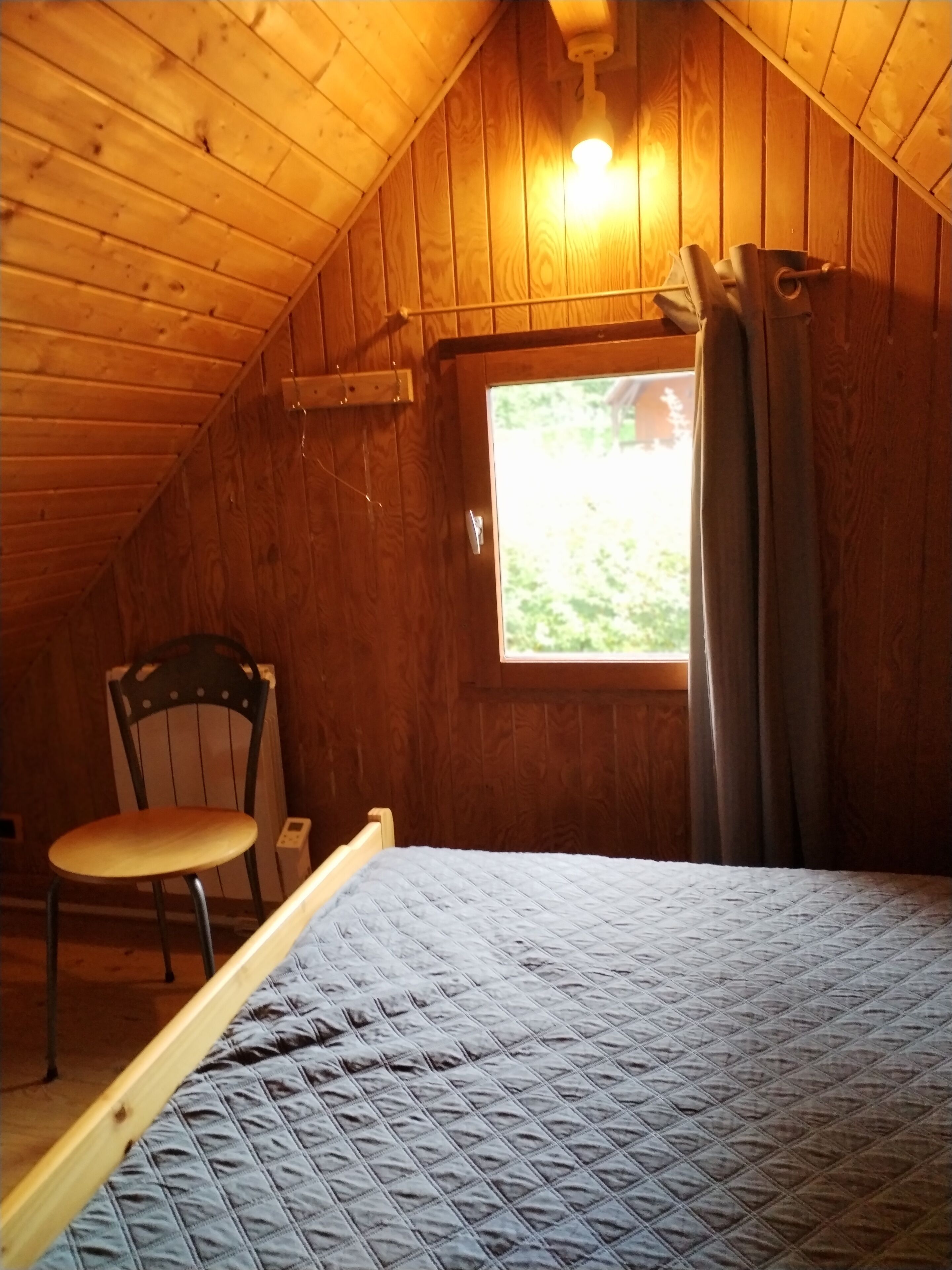 Un pequeño y acogedor dormitorio en el ático con techo inclinado, una cama individual, una silla, un ventanal con cortinas y un radiador.