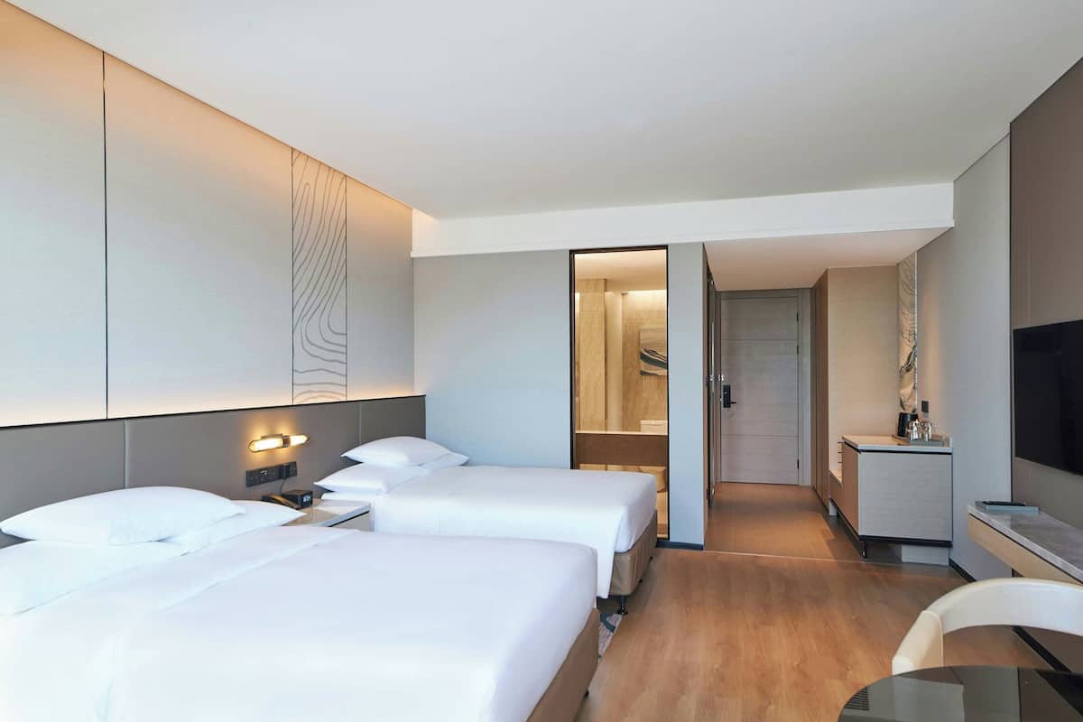 Ein modernes Hotelzimmer mit zwei Betten, einem Schreibtisch, einem Fernseher und einem durch eine offene Tür einsehbaren Badezimmer.