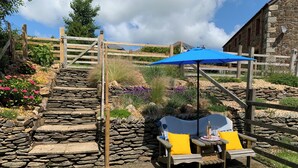 Una escalera de piedra que conduce a un portón de madera, un muro de piedra y un área de descanso con una sombrilla azul y cojines amarillos.