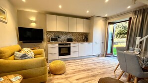 Una sala de estar moderna con un sofá, un reposapiés y una mesa de centro. Se observa una cocina con armarios y un microondas. Un televisor está montado en la pared.