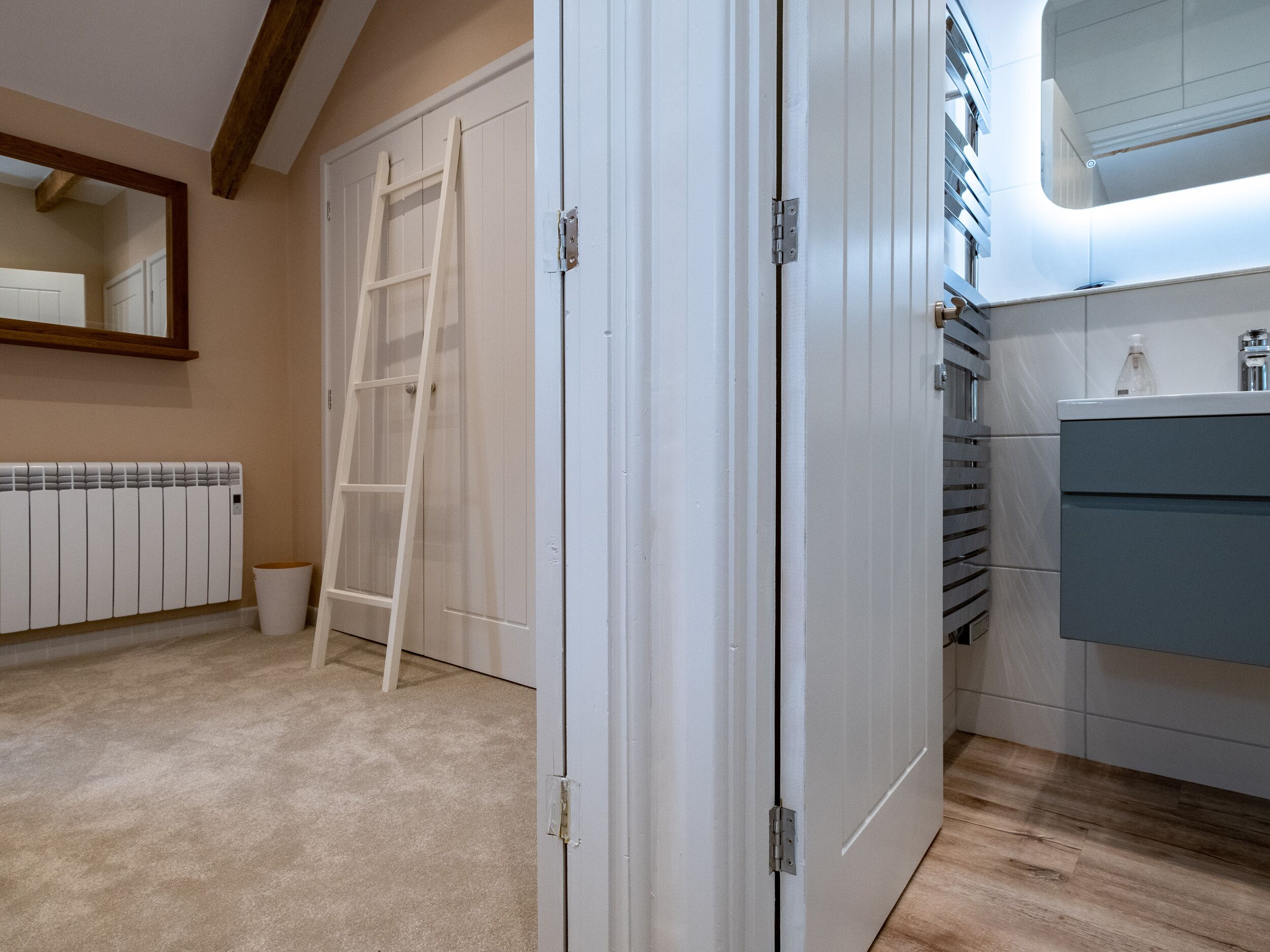A bathroom with a white door, a white radiator, a wooden ladder, and a sink with a mirror above it.