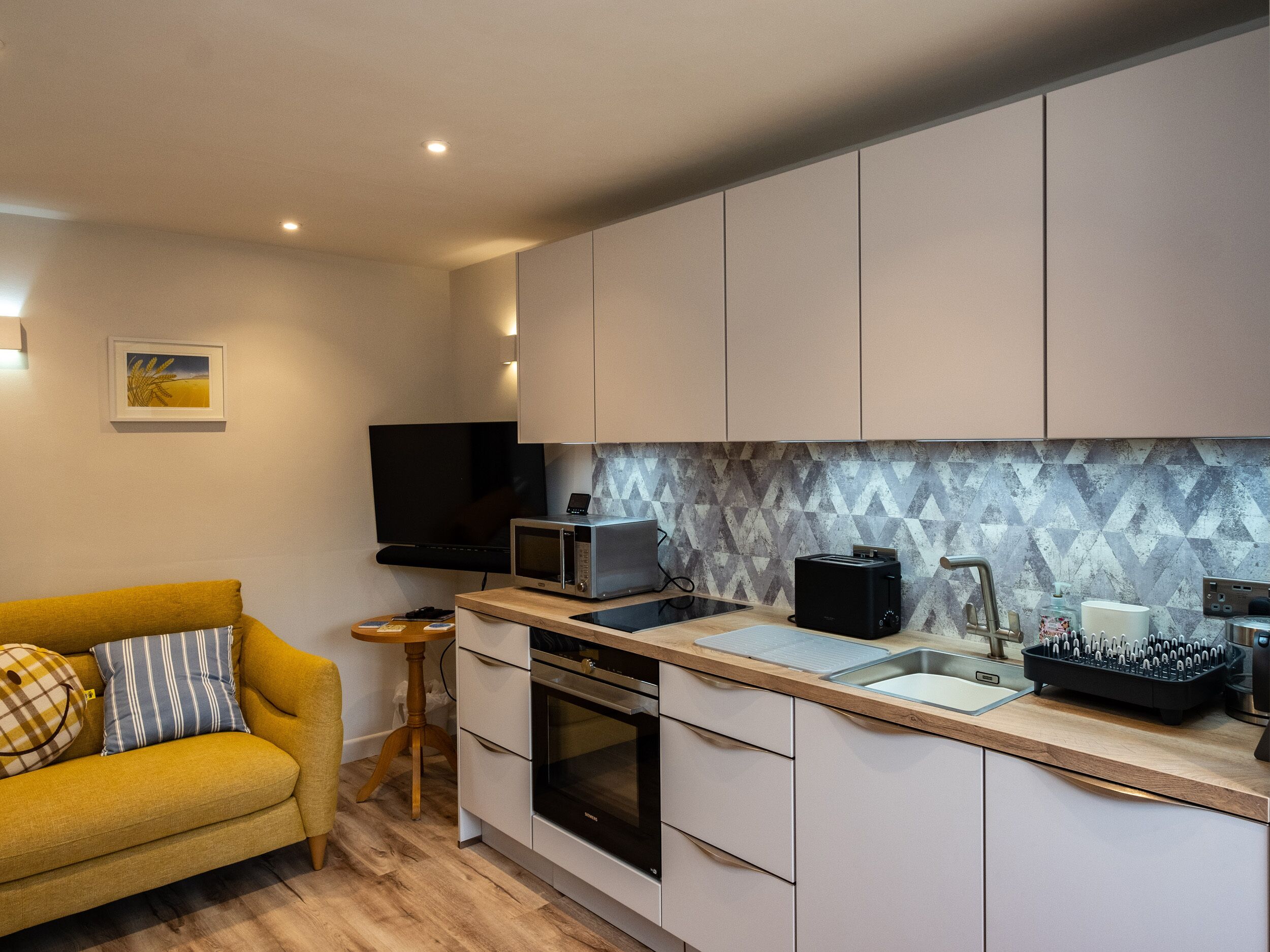 A modern kitchen with white cabinets, a microwave, toaster, and a sink. There's a yellow sofa with striped pillows and a small wooden stool.