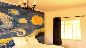 Un dormitorio con un cuadro grande de un cielo nocturno en la pared, una cama con ropa de cama blanca, una araña y una ventana con cortinas.