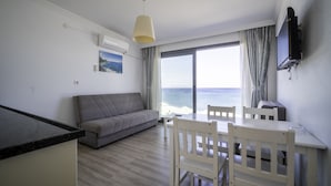 Uma sala de estar moderna com um sofá cinzento, uma mesa de jantar com cadeiras, uma televisão na parede e uma janela grande com vista para o mar.