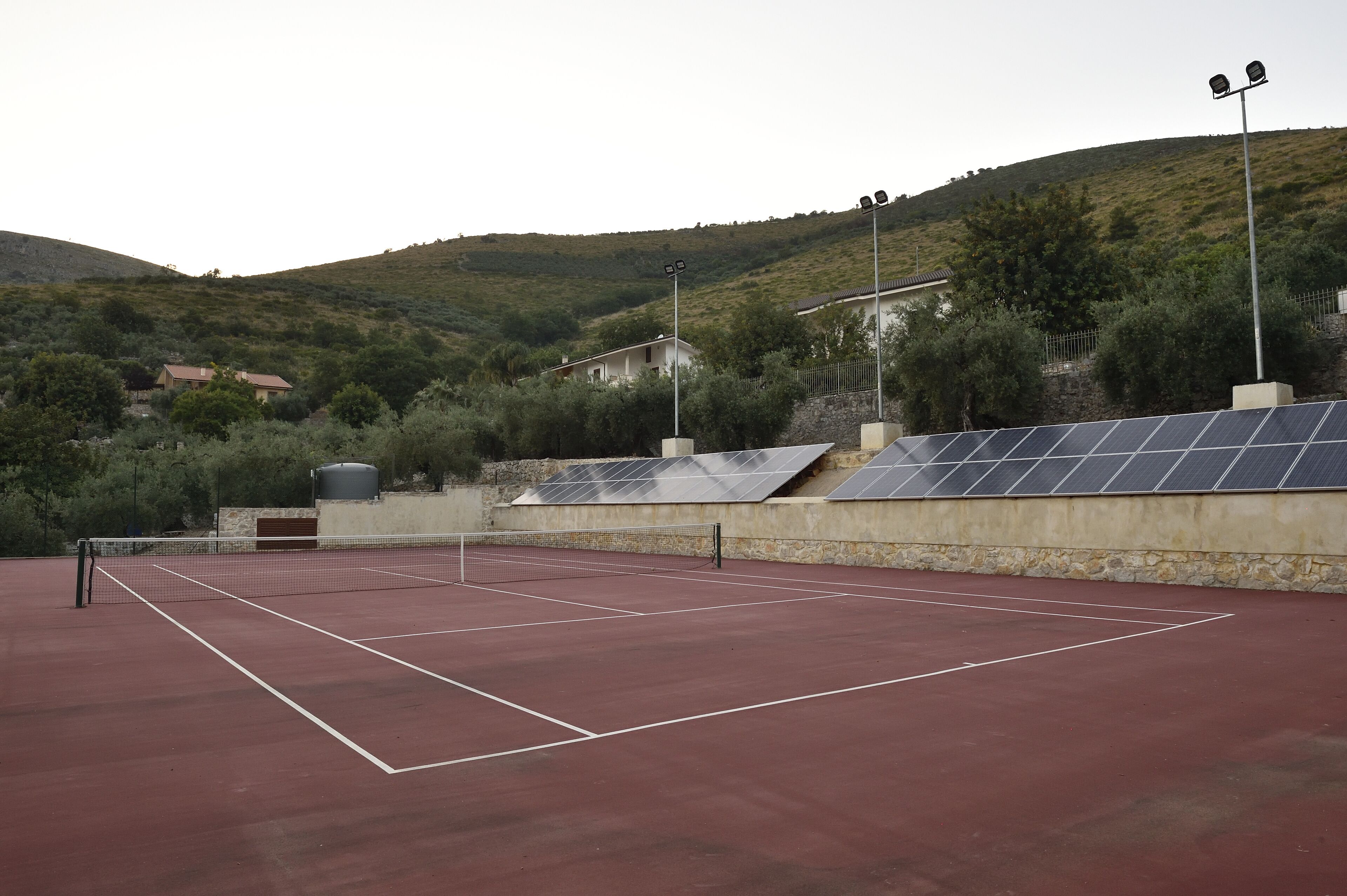 A tennis court with a net, surrounded by stone walls and solar panels.