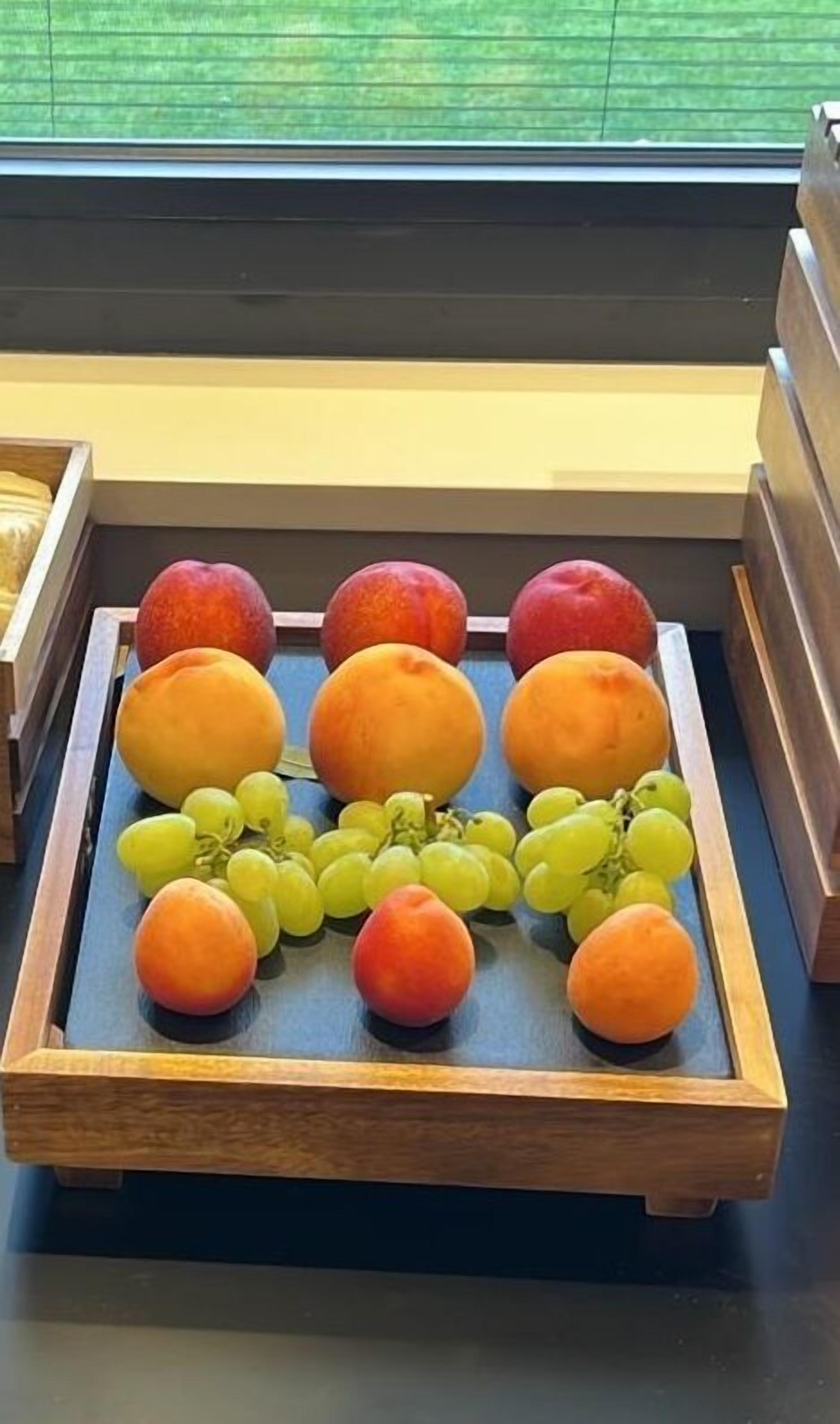 A wooden fruit tray with peaches, grapes, and an orange on a table.