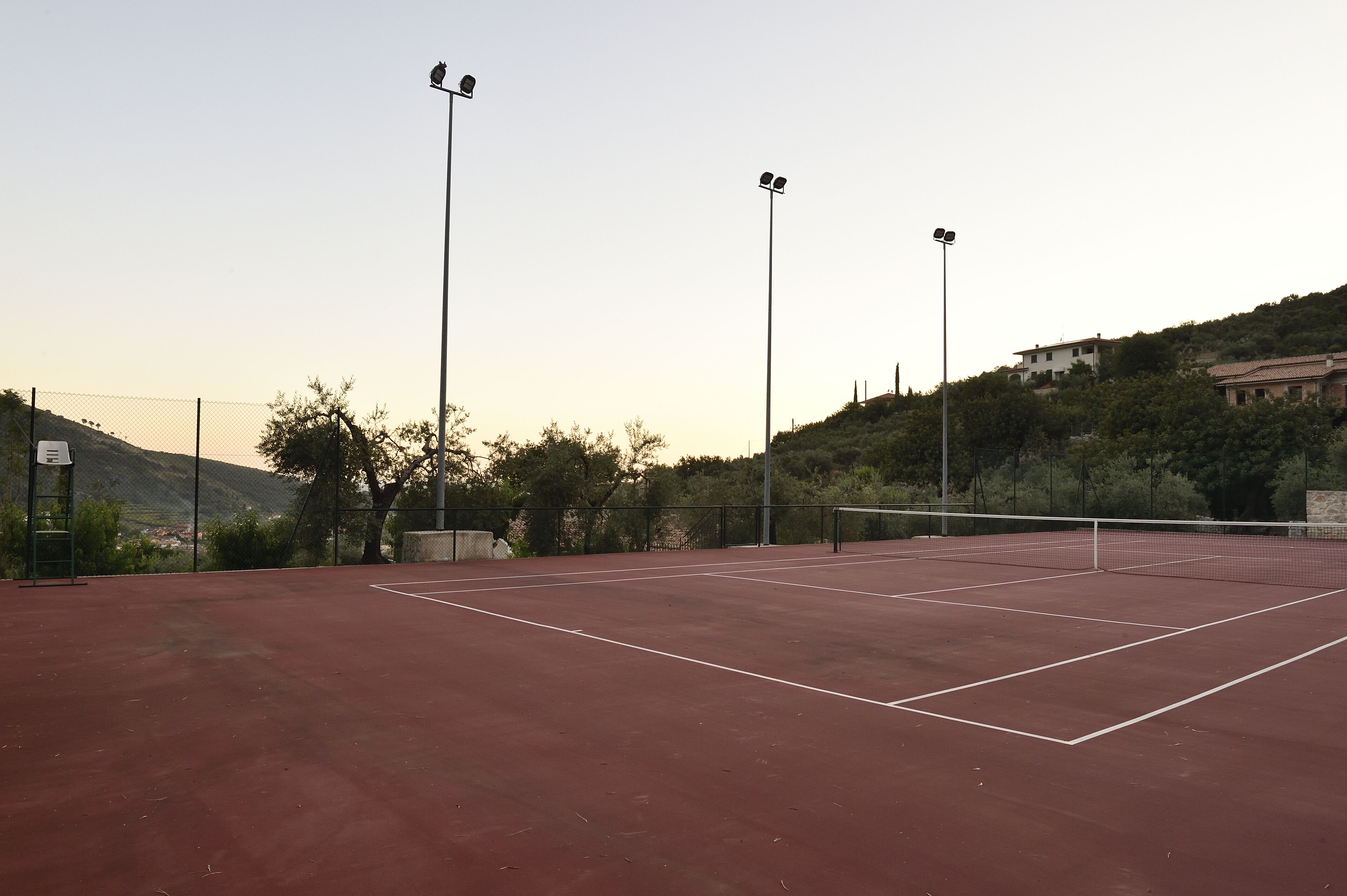 Un court de tennis extérieur, équipé de projecteurs pour les matchs en soirée, entouré d’arbres et situé dans un paysage vallonné.