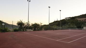 Un court de tennis extérieur, équipé de projecteurs pour les matchs en soirée, entouré d’arbres et situé dans un paysage vallonné.