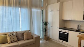 A modern living room with a beige sofa, a small wooden table, and a kitchen area with white cabinets and a black countertop.