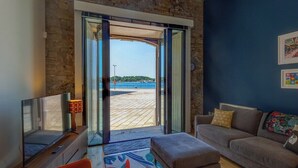 A living room with a TV, a sofa, and a view of a beach through a glass door. - 5 Brewhouse by Pureserviced (Plymouth)