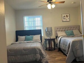 A bedroom with two beds, a ceiling fan, a window with blinds, a bedside table with a lamp, and a framed picture on the wall.