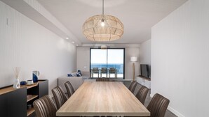 Et moderne spiseområde med et langt træbord og komfortable stole, en stor vævet hængelys og havudsigt gennem store vinduer.