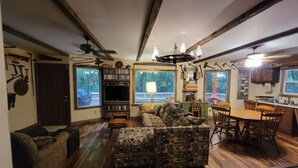 A cozy living room with a patterned sofa, wooden flooring, a bookshelf, and a dining area with chairs and a table. - Charming lake cabin located next to Racoon State Park. (Rockville)