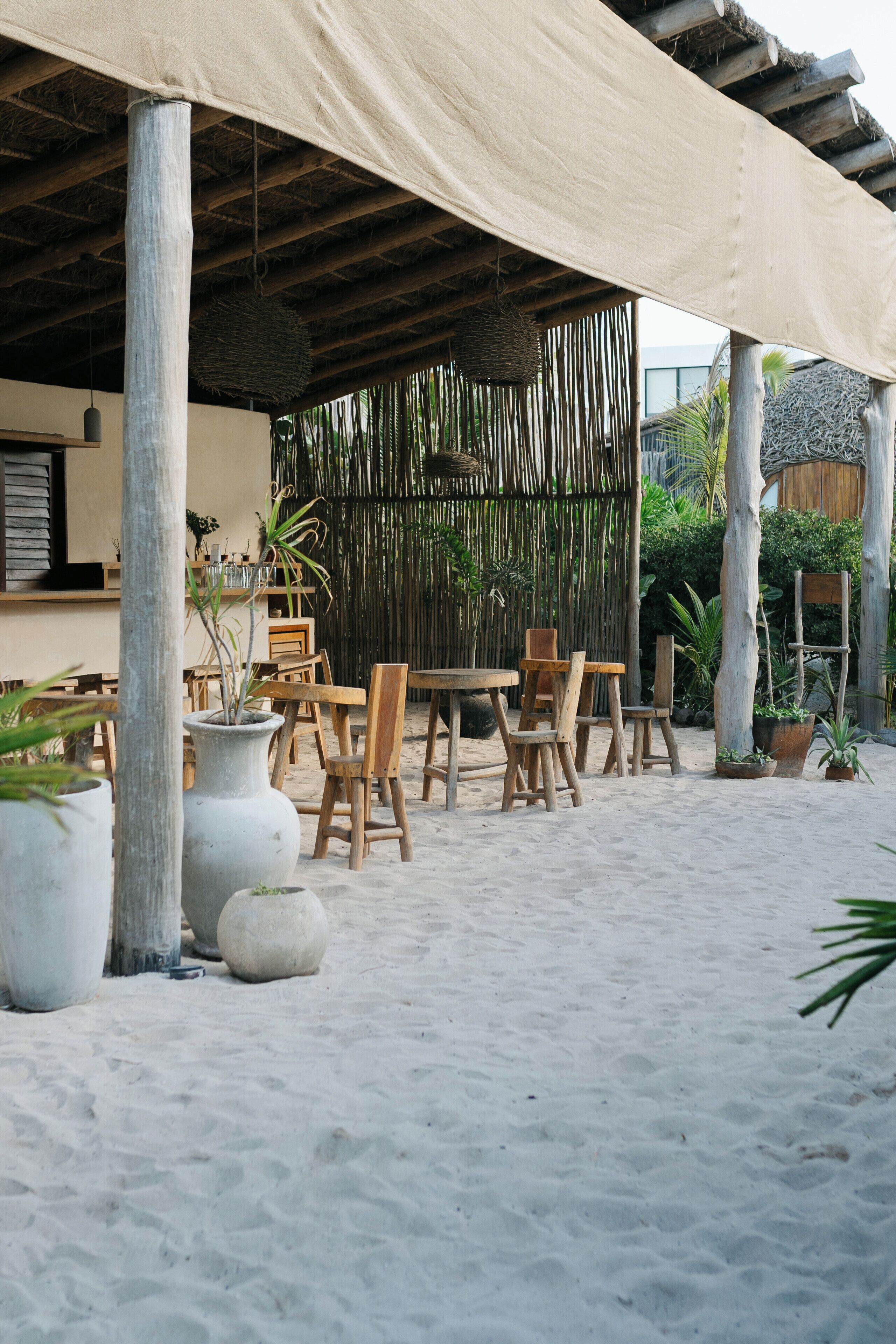 An outdoor seating area with wooden tables and chairs, surrounded by potted plants and a thatched roof structure.
