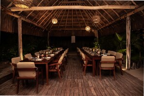 An outdoor dining area with wooden tables and chairs, a thatched roof, and hanging lights.