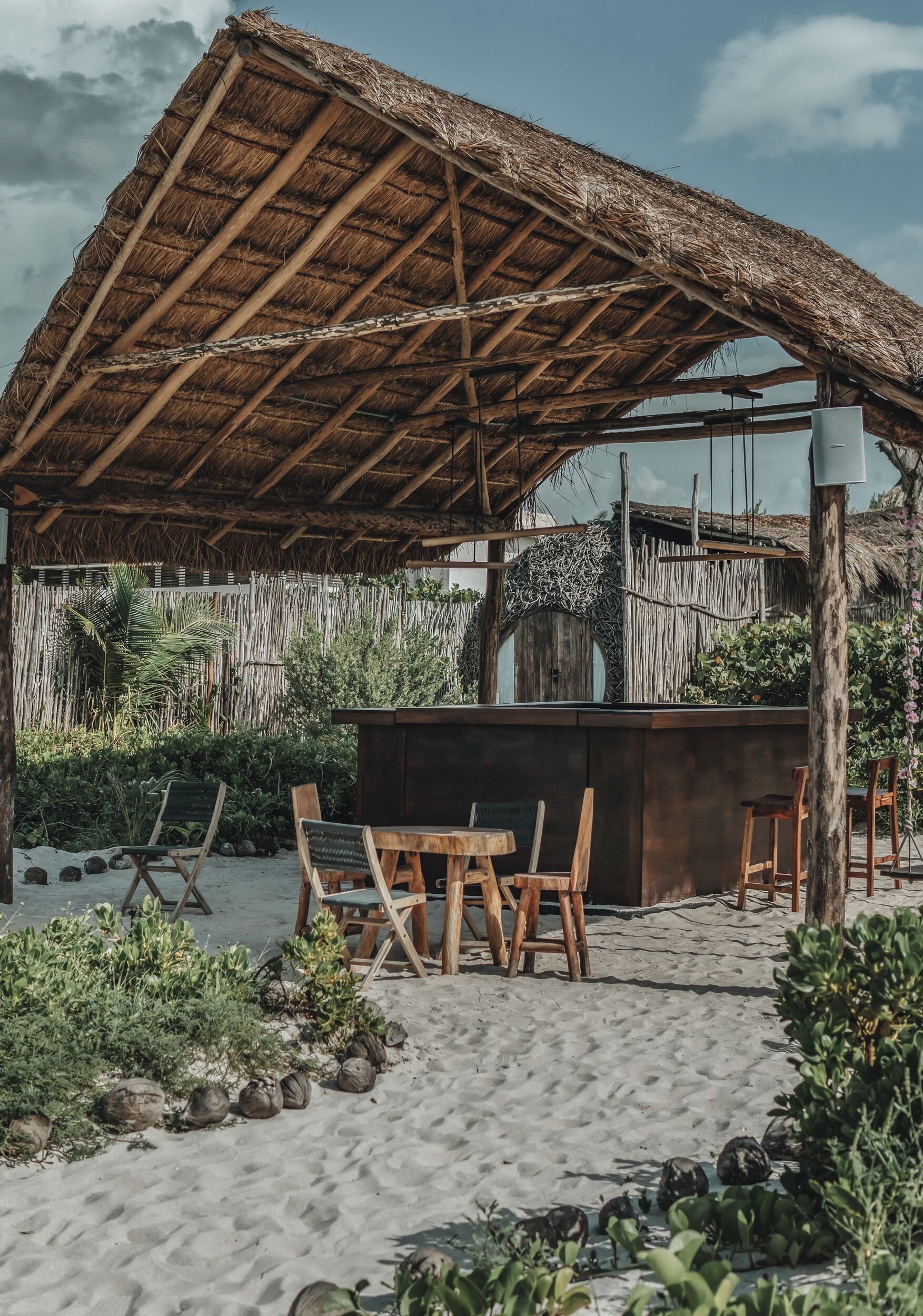 Un área de estar al aire libre frente a la playa, con sillas y una mesa de madera, una estructura de techo de paja y suelo de arena.