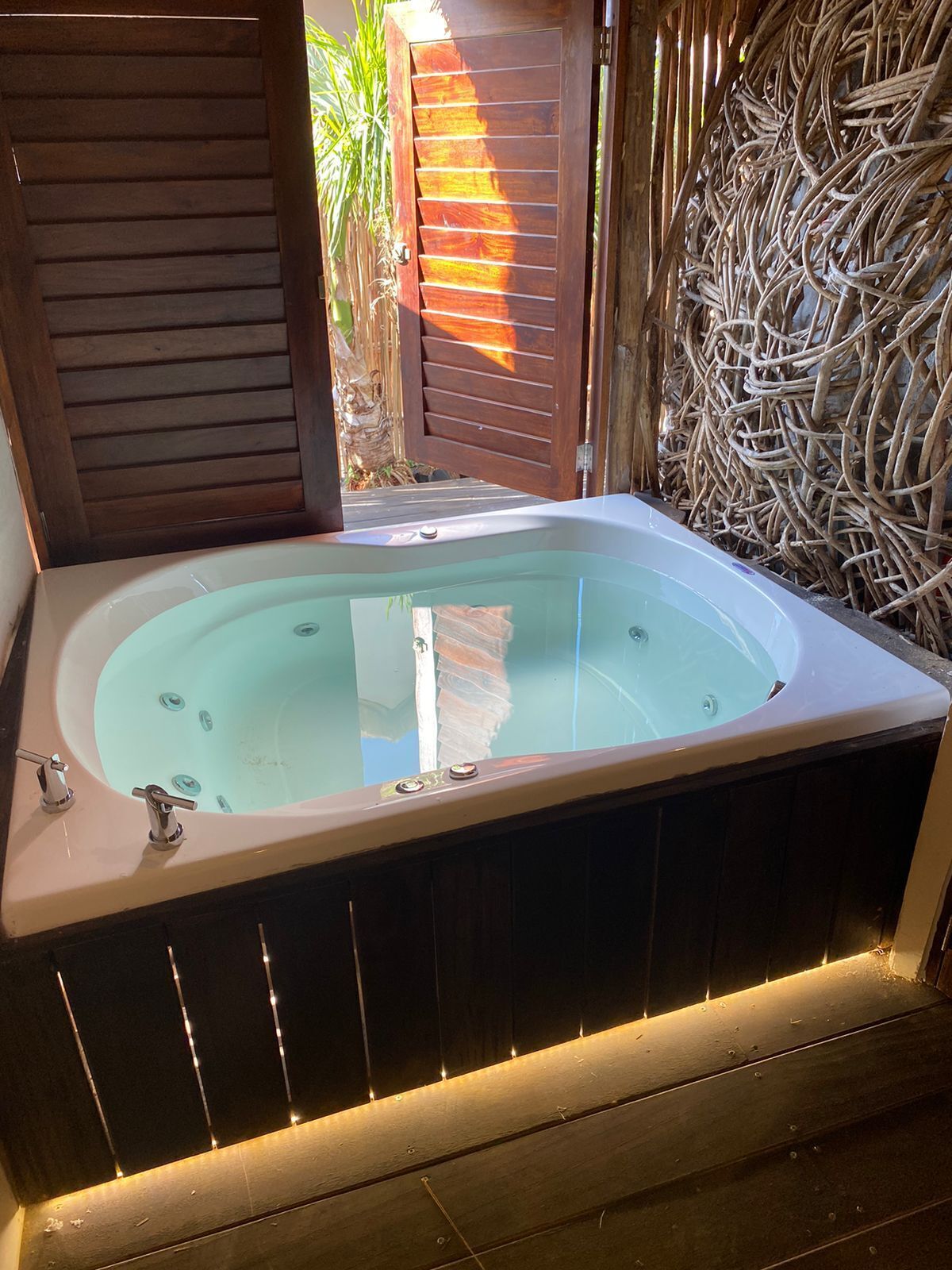 A hot tub with a wooden frame and a glass enclosure, surrounded by a wooden deck and a woven wall.
