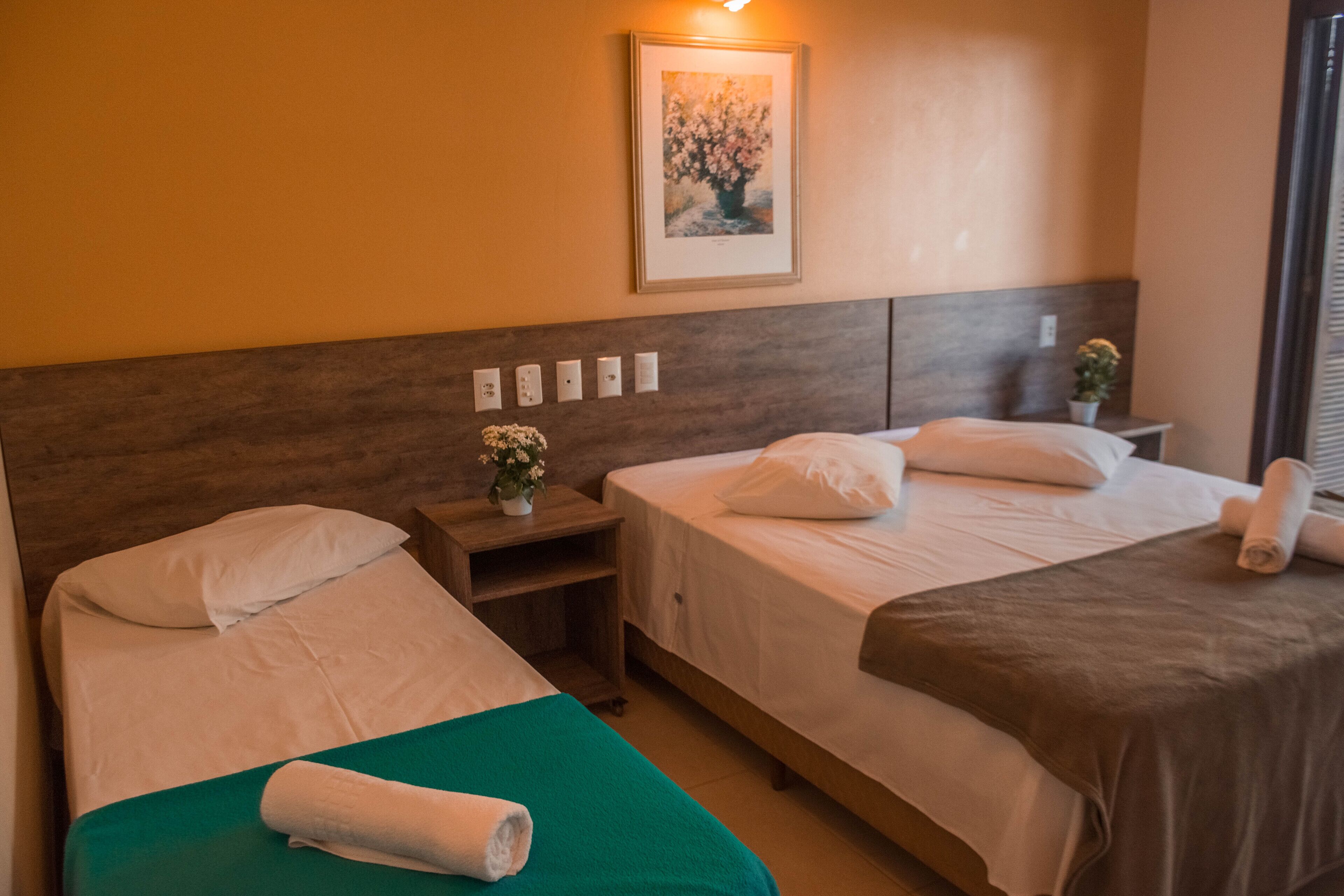 A hotel room with two beds, a wooden headboard, a small bedside table with a vase of flowers, and a framed picture on the wall.