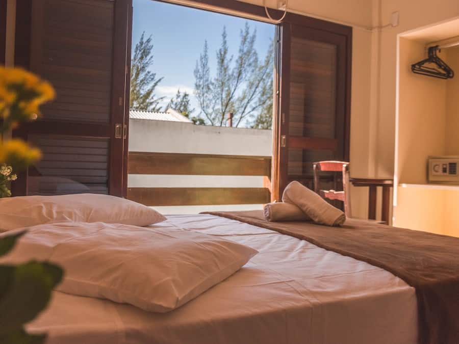 Un dormitorio con una cama grande, una mesita y una silla. Se ve una ventana con vistas a árboles y un edificio.
