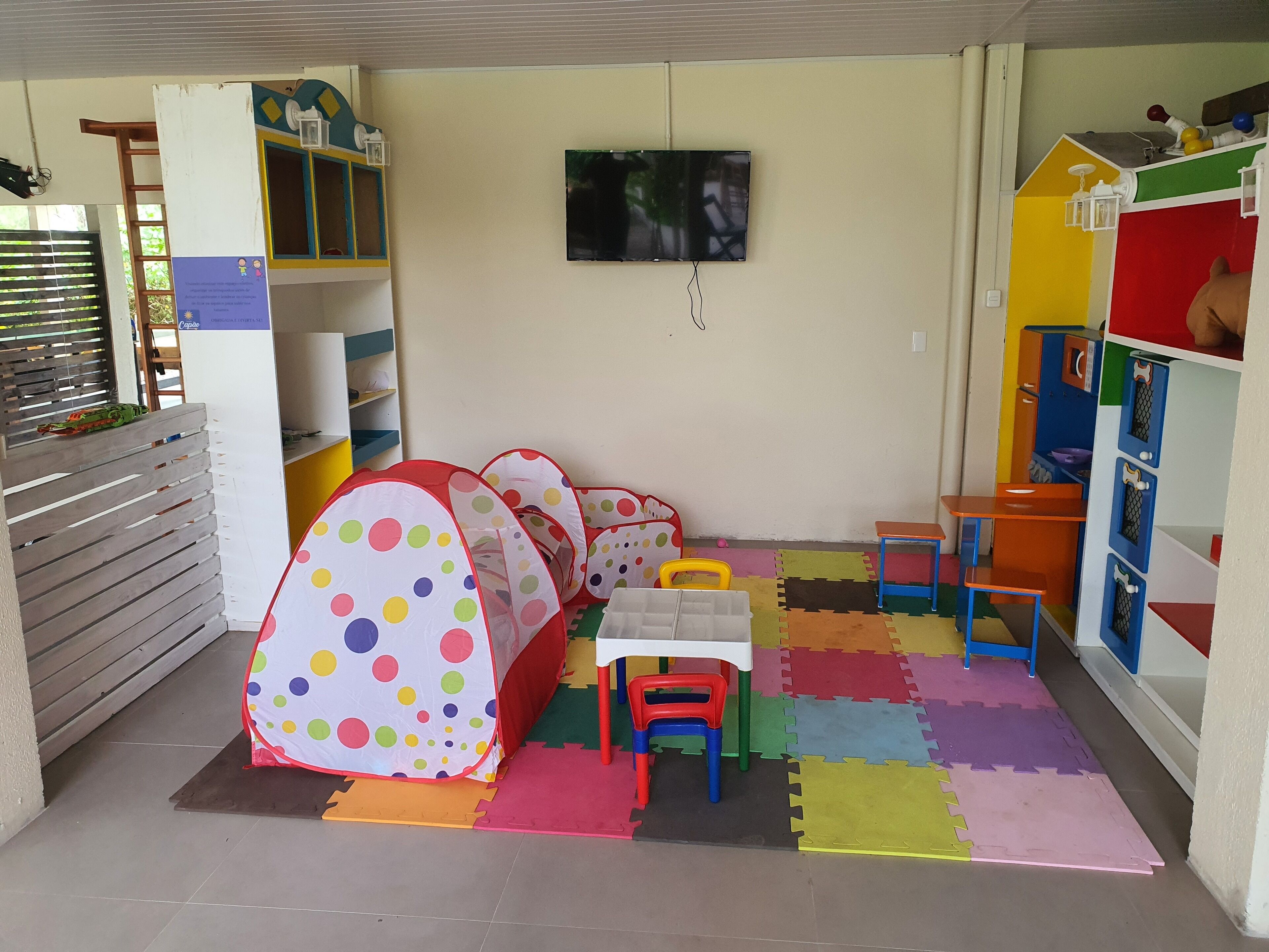 A children's playroom with colorful play tents, a small table, and a TV mounted on the wall.