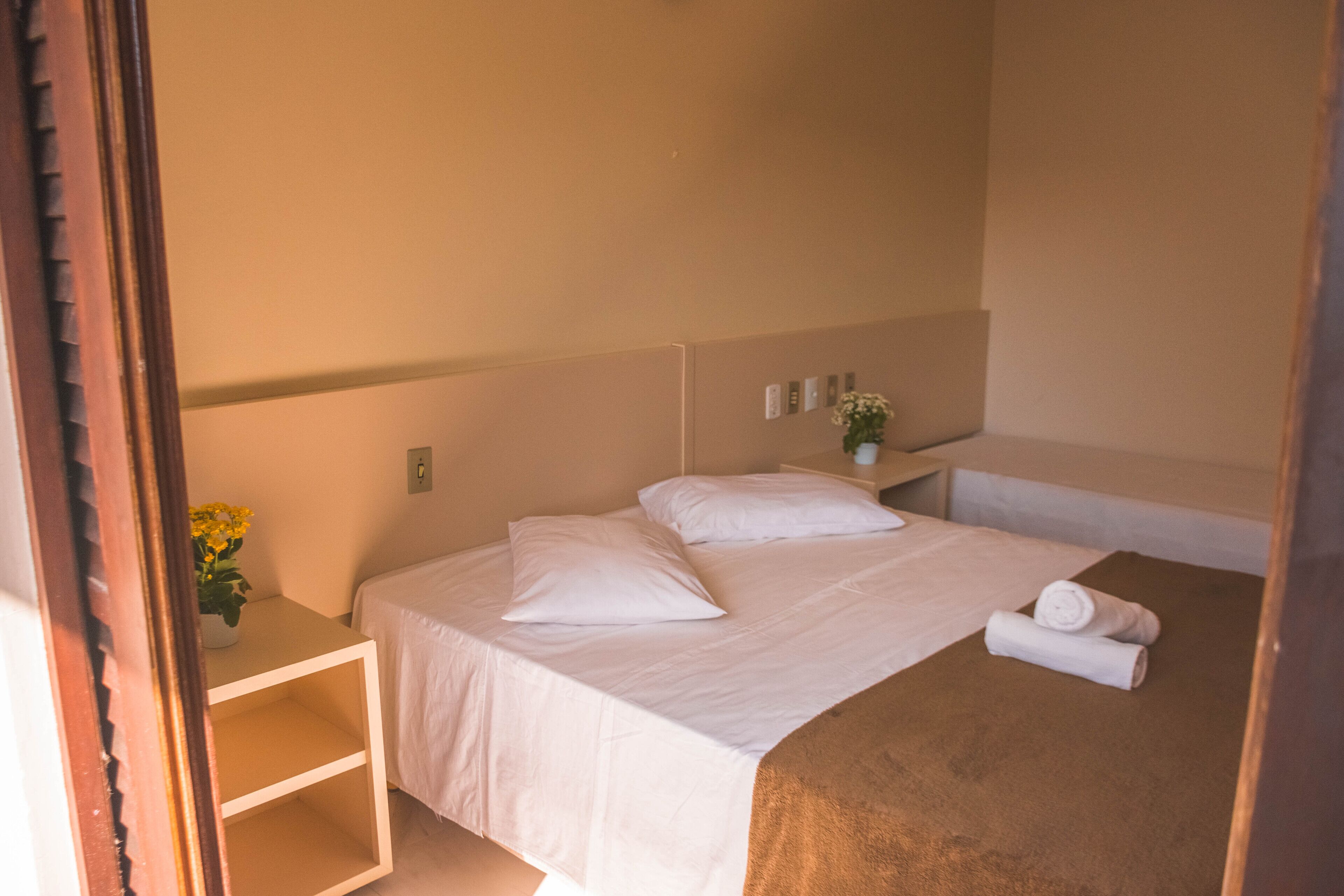 A neatly made bed with white linens and pillows, a small bedside table with a vase of flowers, and a brown blanket on the bed.