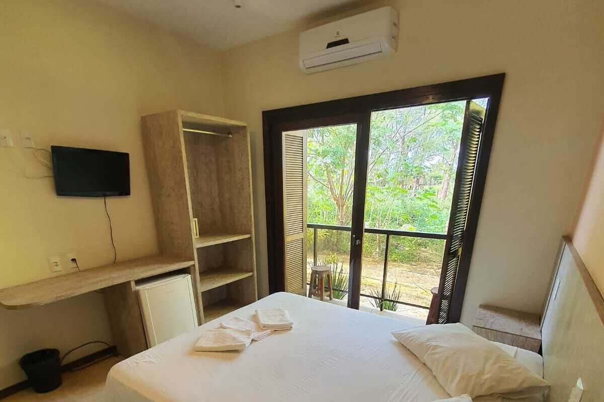 A bedroom with a bed, a desk, a TV, a wardrobe, and a window with a view of greenery.