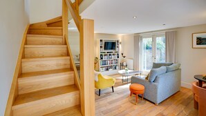 A modern living room with a wooden staircase, a sofa, a coffee table, and a bookshelf. - South Stable: Cosy Stone Retreat with Parking (Alnmouth)