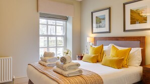 A neatly made bed with yellow pillows and a beige quilt, a window with a view, and a framed picture on the wall. - Pope Lodge: Cosy Stone Coach House in Alnmouth (Alnmouth)