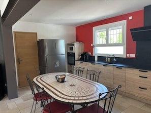 A modern kitchen with a tiled dining table, stainless steel appliances, and a red accent wall. - Christine's Cabanon (L'Isle-sur-la-Sorgue)