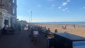 A beachside promenade with people walking and sitting, overlooking a sandy beach and the sea.