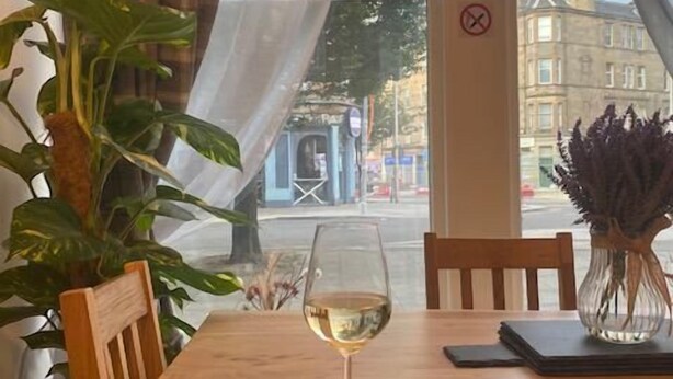 A wooden table with a glass of white wine, a black notebook, and a vase with purple flowers.