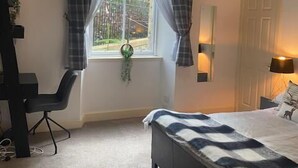 A bedroom with a bed, a desk, a chair, a window with curtains, a hanging plant, and a lamp.