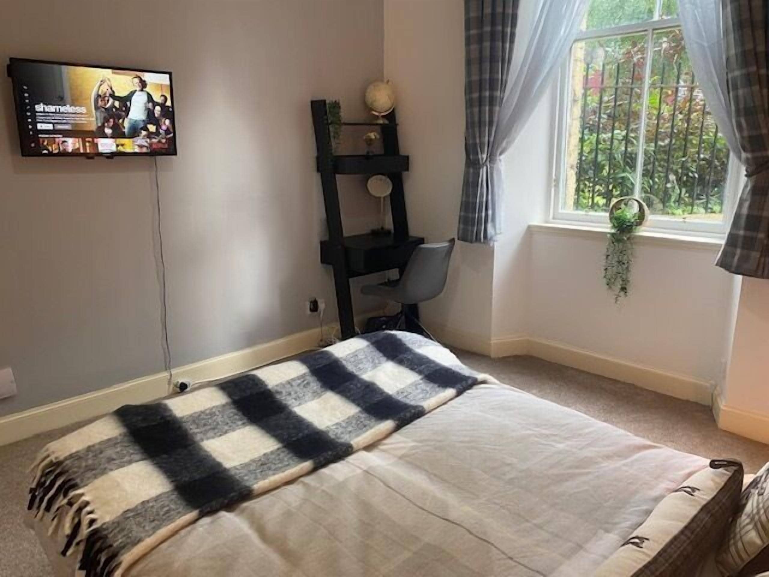Un dormitorio con cama, televisor montado en la pared, escritorio y ventana con cortinas.