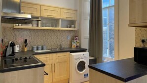A compact kitchen with wooden cabinets, a built-in oven, a washing machine, and a window with curtains.