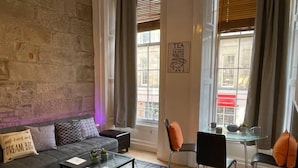 A cozy living room with a stone wall, a sofa with patterned cushions, a glass coffee table, and a view of the street through large windows.