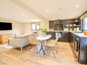 A modern kitchen with a dining area, wooden flooring, and a flat-screen TV mounted on the wall. - The Cabin at Evelyns Retreat (Corwen)