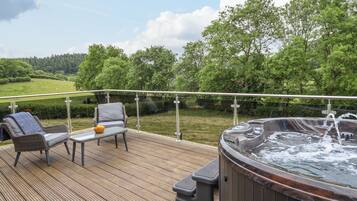Une terrasse en bois avec des meubles d’extérieur, un jacuzzi et une vue panoramique sur un paysage verdoyant, avec des arbres et un champ.