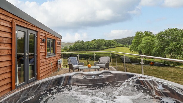 A wooden cabin with a hot tub, two chairs, and a table on a deck overlooking a green landscape. - The Cabin at Evelyns Retreat (Corwen)