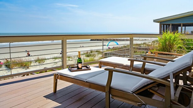 A wooden deck with lounge chairs and a bottle of champagne, overlooking a beach with people and a lifeguard tower.