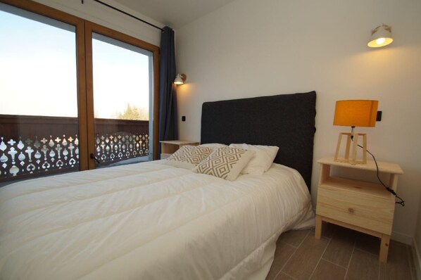Une chambre à coucher avec un grand lit, une table de chevet avec une lampe et une vue sur l’extérieur, visible à travers une fenêtre agrémentée d’un paravent décoratif.