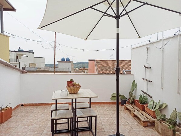 A rooftop terrace with a table and chairs, surrounded by potted plants and string lights.