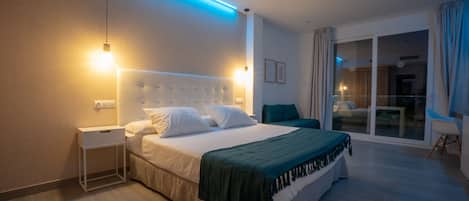 A modern hotel room with a large bed, a bedside table, a nightstand, a green throw blanket, and a view of the outside through a sliding glass door.