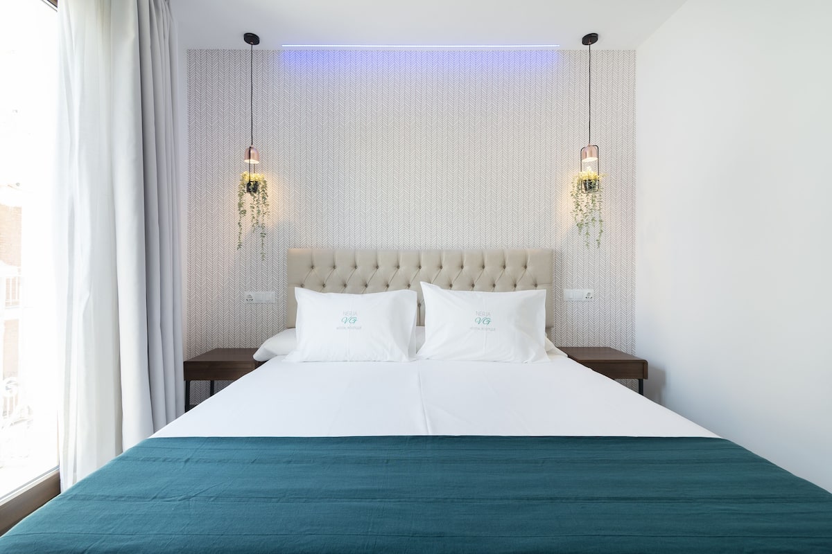 Una cama bien hecha con una colcha verde azulada, dos almohadas blancas y un cabecero capitonado. Hay dos mesitas de noche con lámparas, y la pared presenta un patrón de espiga.